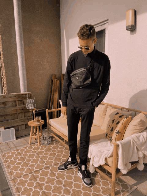 a man wearing a black sweatshirt and black pants stands in front of a wooden couch