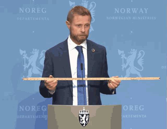 a man in a suit and tie stands at a podium holding a tape measure in front of a sign that says norway
