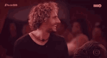 a man with curly hair is smiling while standing in front of a crowd of people .