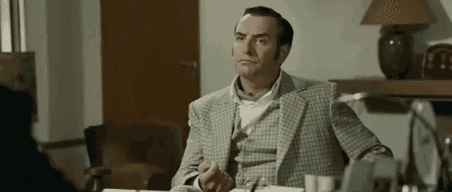 a man in a suit is sitting at a desk in a room with a lamp .