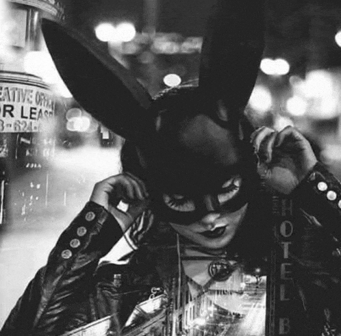 a woman wearing a bunny mask and a leather jacket is standing in front of a building .