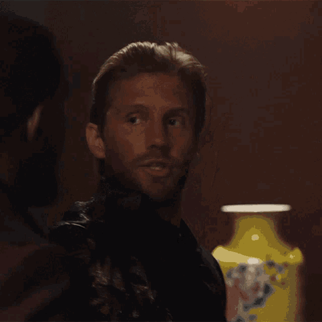 a man with a beard looks at another man in front of a yellow vase