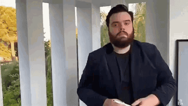 a man with a beard wearing a suit and tie is standing in front of a window .