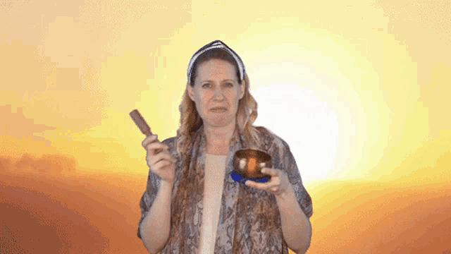 a woman is holding a bowl and a wooden stick in front of a sunset