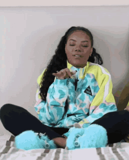 a woman is sitting on a bed wearing blue slippers and a colorful jacket