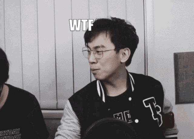 a man wearing glasses and a varsity jacket is sitting in front of a microphone and saying wtf .