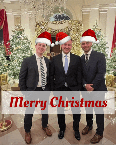three men wearing santa hats are standing in front of christmas trees and a merry christmas banner