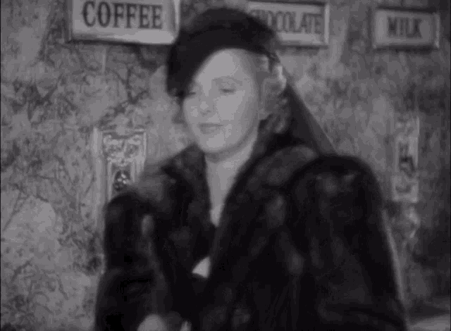 a woman wearing a fur coat is standing in front of a wall with signs for coffee chocolate and milk
