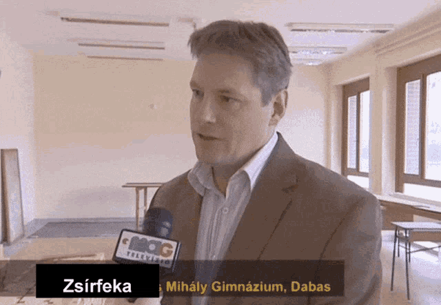 a man in a suit is talking into a microphone with the name zsirfeka behind him