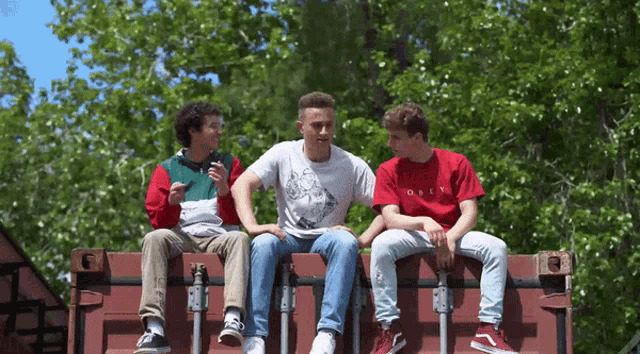 three young men are sitting on top of a red container with one wearing a red shirt that says obey