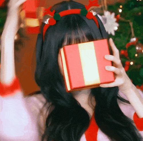 a woman wearing a reindeer headband is holding a christmas present in front of her face