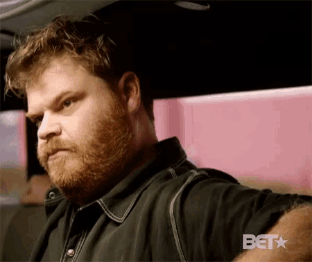 a man with a beard is sitting in a car with a bet logo in the corner .