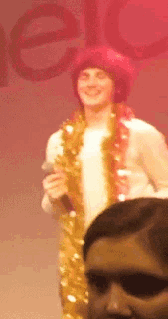 a man wearing a tinsel necklace holds a microphone and smiles