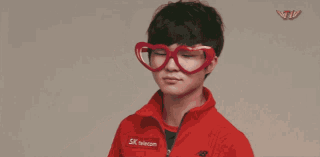 a young man wearing heart shaped glasses and a red jacket makes a heart shape with his hands