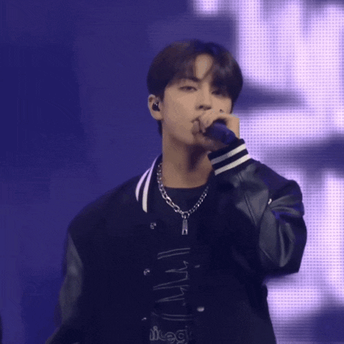 a man in a black jacket is singing into a microphone while wearing a necklace with the letter l on it
