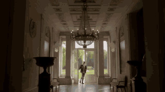 a man in a suit is standing in a hallway with a chandelier hanging from the ceiling