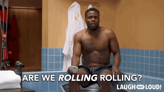 a shirtless man is sitting on a toilet with the words are we rolling rolling laugh out loud behind him