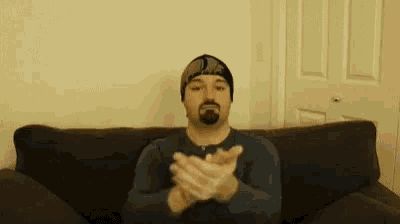 a man with a beard is sitting on a couch with his hands together .