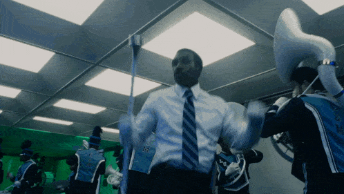 a man in a suit and tie is dancing in front of a marching band with the letter m on their uniform