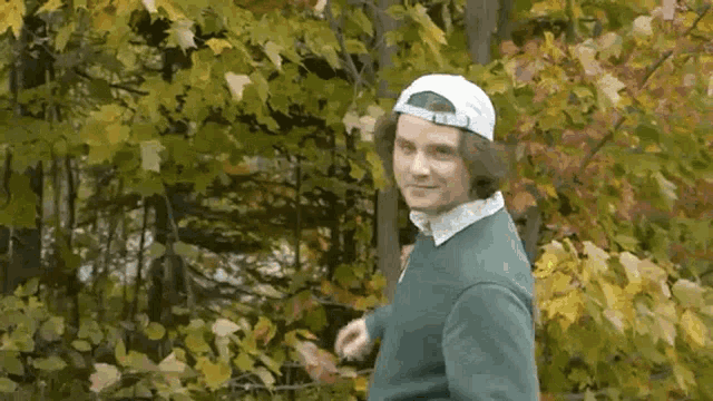 a man wearing a white hat and a green sweater is standing in front of a tree .