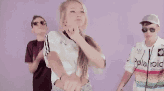 two men and a woman are dancing in front of a purple background and one of the men is wearing a polaroid shirt