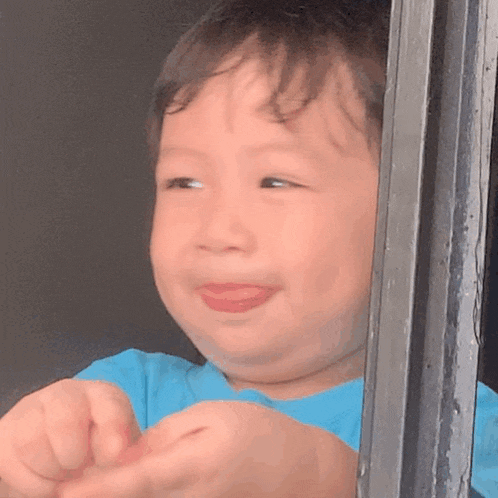 a baby in a blue shirt is looking out of a window and making a funny face .