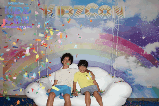 two young boys are sitting on a cloud shaped couch in front of a kidzcon sign