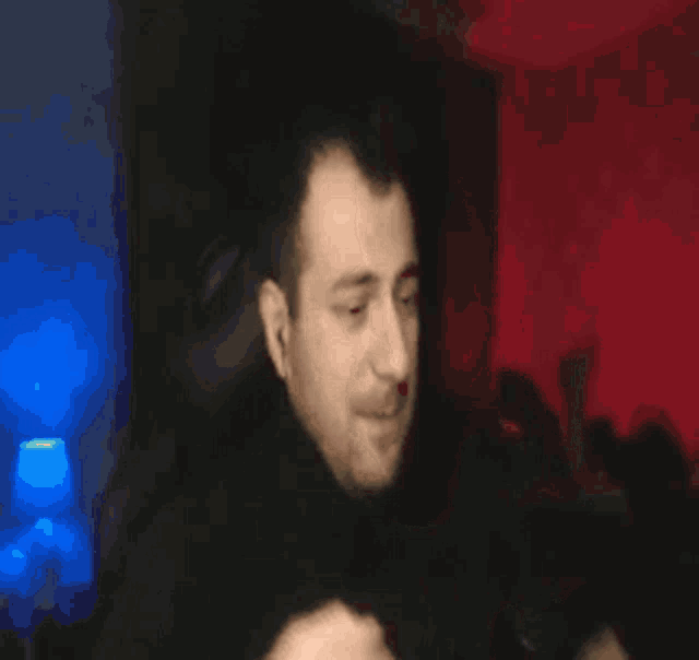 a man with a beard is sitting in a dark room with a red curtain behind him
