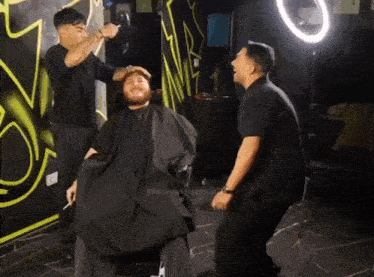 a man in a black cape is getting his hair cut by two barbers