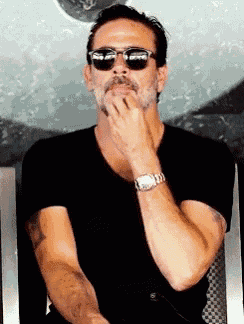 a man wearing sunglasses and a black shirt has his hand on his chin .