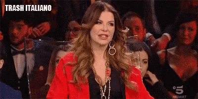 a woman in a red jacket is holding a glass of wine in front of a crowd of people and says trash italiano .