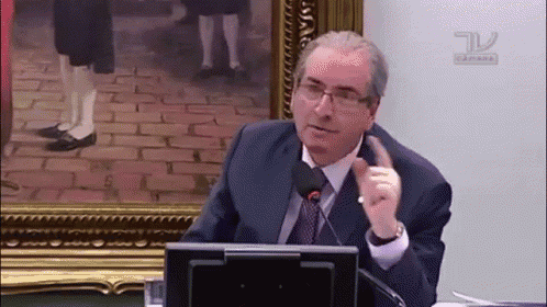 a man in a suit and tie speaking into a microphone in front of a framed picture with the letters tv on it