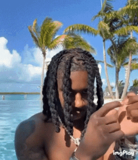 a man with dreadlocks is smoking a cigarette in front of a pool with palm trees in the background .
