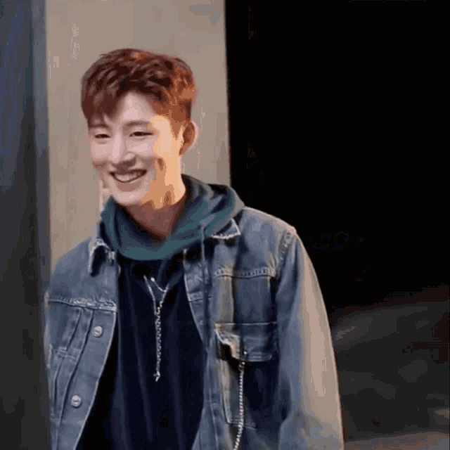 a man wearing a denim jacket and a green hoodie smiles