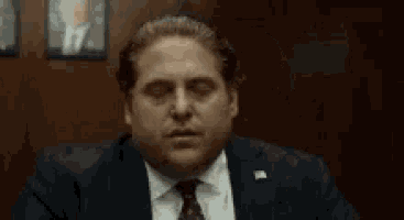 a man in a suit and tie is sitting at a table with his eyes closed and his hand up .