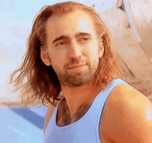 a man with long hair and a beard wearing a blue tank top is looking at the camera .