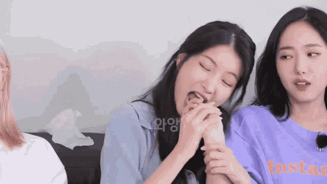 two young women are sitting next to each other on a couch . one of the girls is licking her hand .