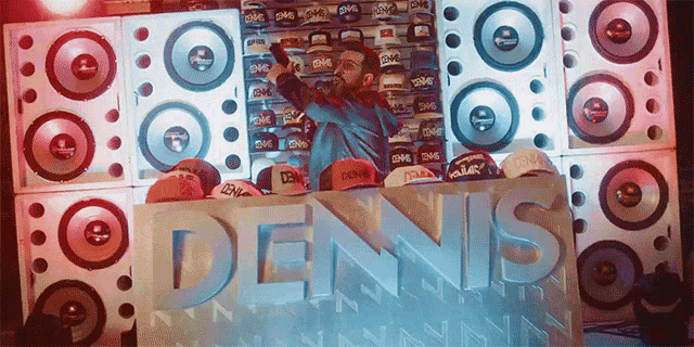 a man stands behind a dennis sign surrounded by speakers