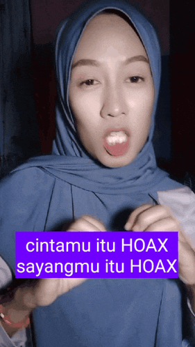 a woman wearing a blue hijab is making a funny face with a purple sign that says cintamu itu hoax