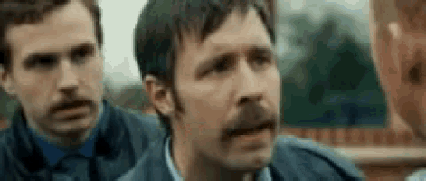 a man with a mustache is talking to another man while another man looks on .