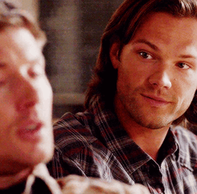 a man with long hair and a plaid shirt looks at another man
