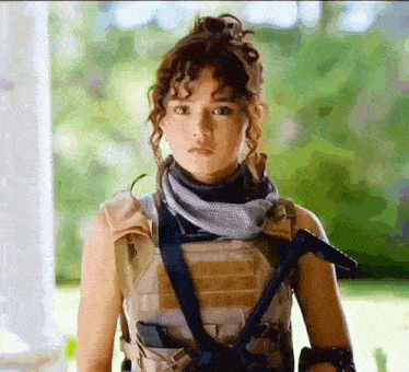 a woman with curly hair is wearing a tactical vest and scarf and holding a gun .