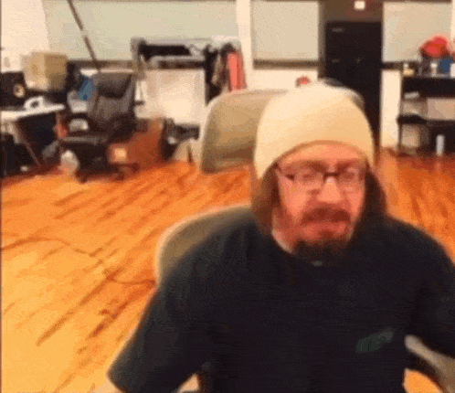a man with a beard and glasses is sitting in a chair in a room with a wooden floor .