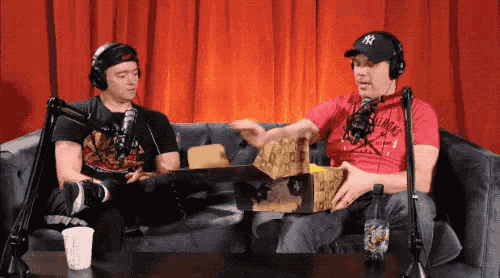 two men are sitting on a couch in front of a red curtain and one is wearing a ny hat