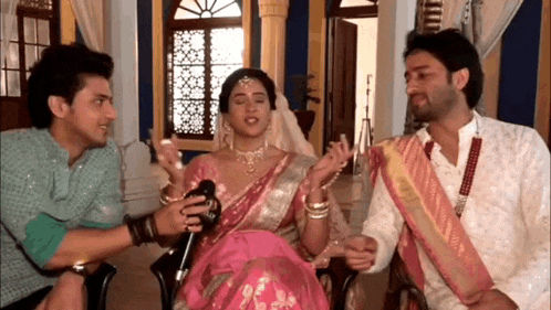 two men and a woman are sitting in a room talking to each other . the woman is wearing a pink saree .
