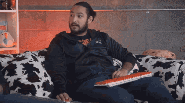 a man sitting on a cow print couch holding a keyboard and wearing a jacket that says snc