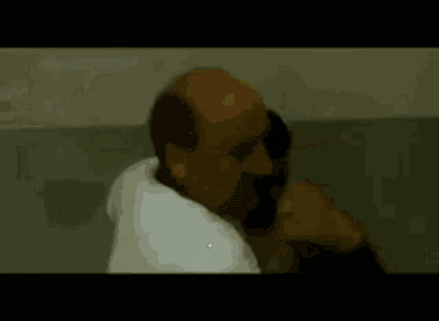 a man in a white shirt is talking on a cell phone while standing next to a wall .