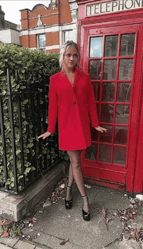 a woman in a red coat is standing in front of a red phone booth .