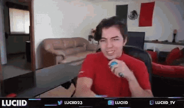 a man in a red shirt is sitting at a desk in a living room holding a remote control .