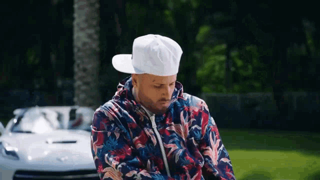a man wearing a hat and a floral jacket is standing in front of a white car .